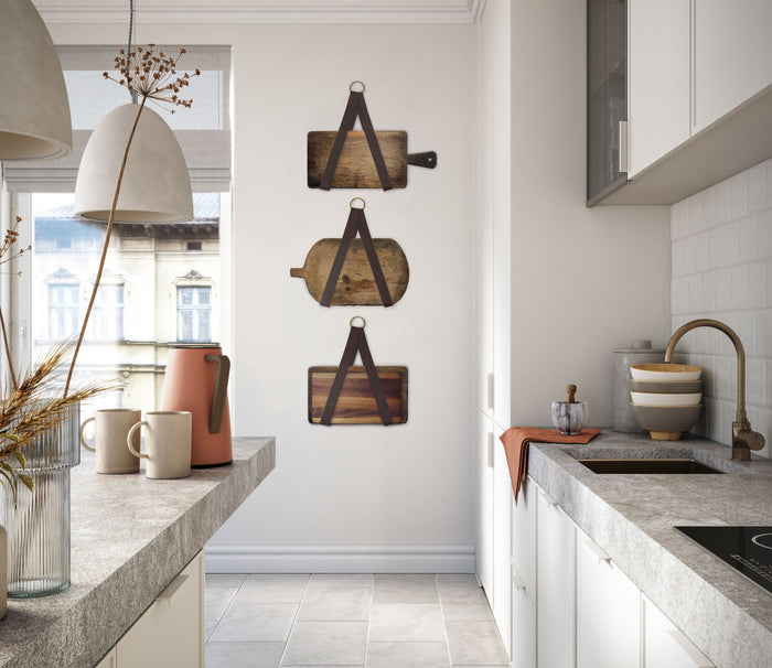 Set of Keyaiira leather display holders styled in a bright modern kitchen, each handcrafted leather strap with brass ring suspending rustic wooden cutting boards as functional wall decor.