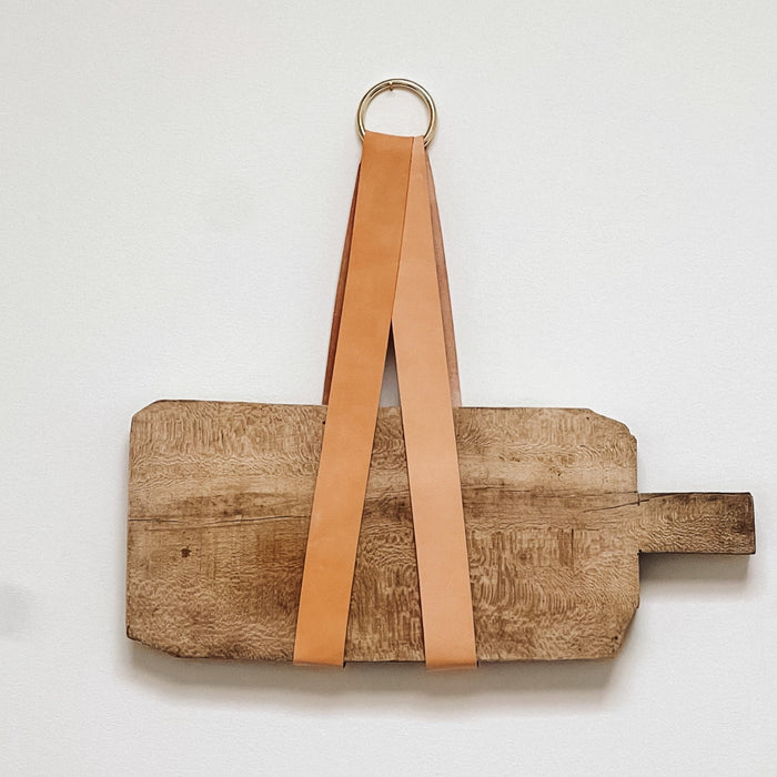Close-up of Keyaiira tan leather display holder with brass ring, mounted on a white wall and holding a vintage wooden cutting board for a natural, minimalist kitchen accent.
