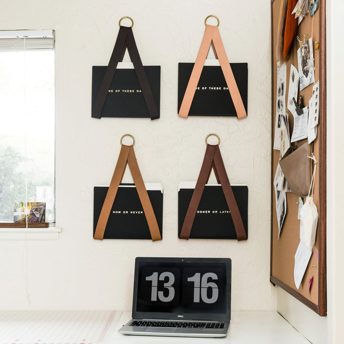 Keyaiira wall-mounted leather file holders in assorted colors and finishes, each handcrafted leather strap with brass ring holding black file folders for modern office organization.