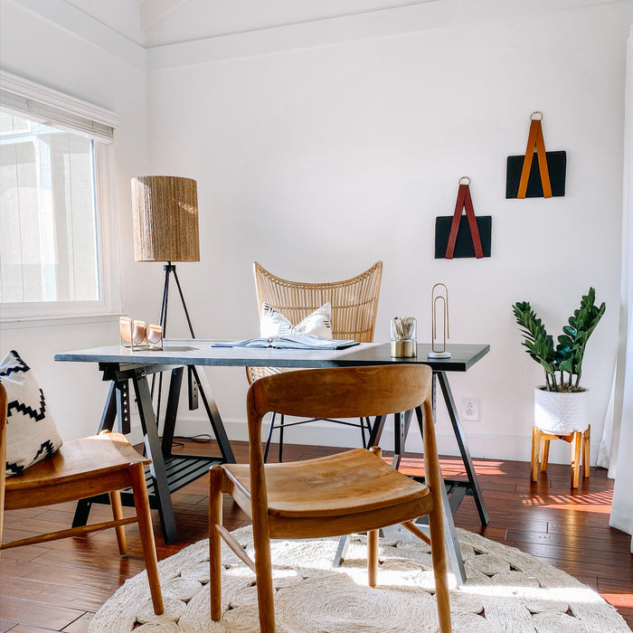 Keyaiira leather file holders displayed on a white office wall — artisan vegetable-tanned leather straps with brass rings holding black folders. Minimal modern workspace styling with wood furniture and natural light.