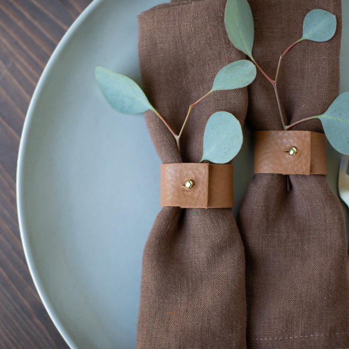 A pair of brown leather napkin rings on a dark napkin, displayed on a plate with some greenery in the background by keyaiira