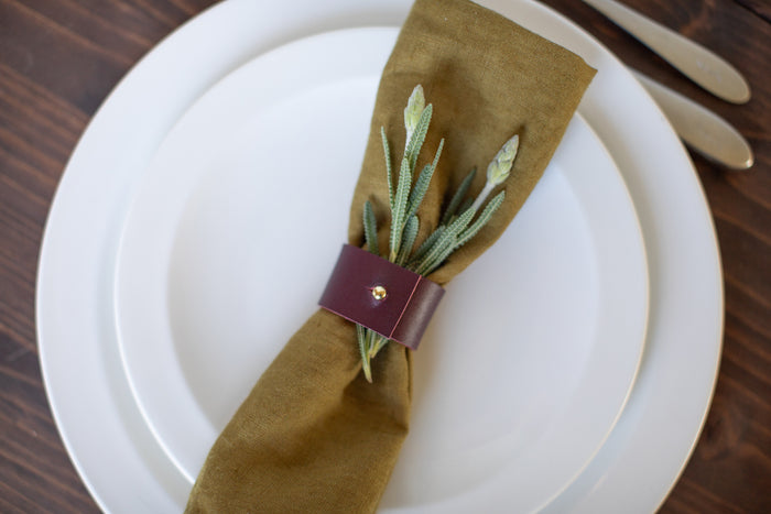 Leather Napkin Ring shown on a white plate with an olive linen napkin and sprigs of greenery  handcrafted leather napkin tie with brass stud adds modern, minimal elegance to table settings by Keyaiira.