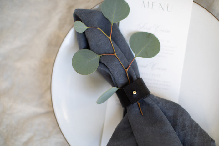Keyaiira Leather Napkin Ring shown with a navy linen napkin, carefully crafted leather tie with brass stud detail adds warm, minimal sophistication to a farmhouse or barn wedding