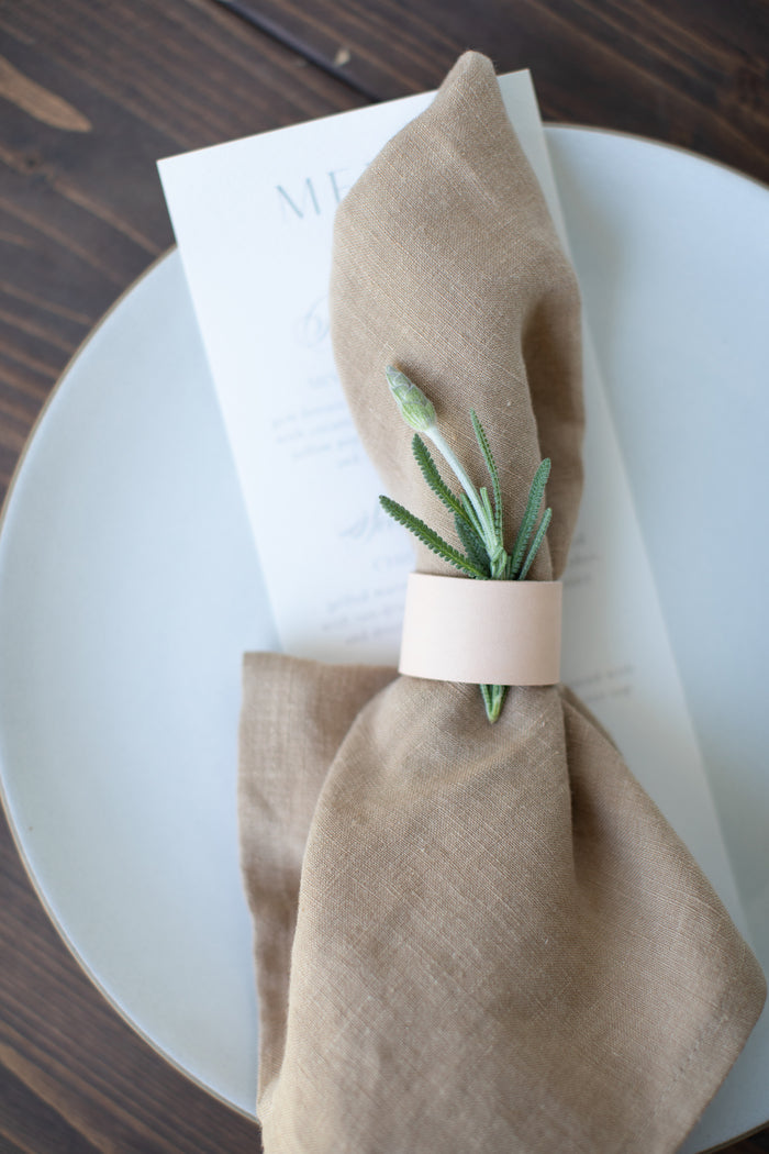 Small-batch crafted Leather Napkin Rings by Keyaiira, genuine leather table accessories with brass hardware shown on a beige plate with linen napkins, perfect for minimal modern dining with menu in the background