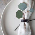 Close-up of a Keyaiira leather napkin tie wrapped around a soft linen napkin, styled on a white ceramic plate with eucalyptus greenery — handcrafted leather napkin holder for minimalist, Scandinavian-inspired table setting and modern dining decor.