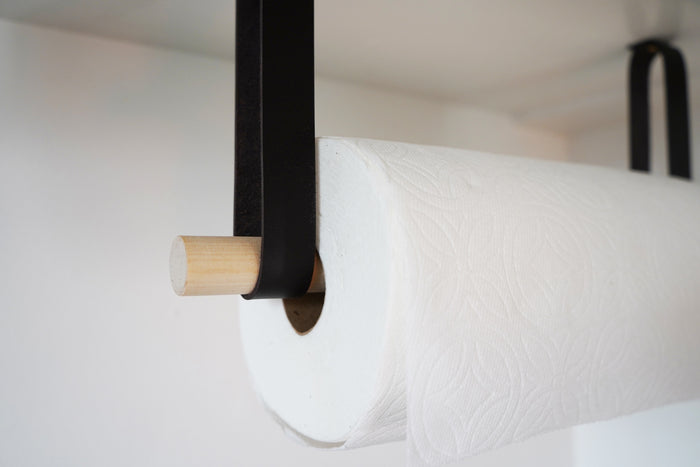 Close-up of black leather wall strap securing wood dowel paper towel rod — minimalist kitchen wall organizer detail.