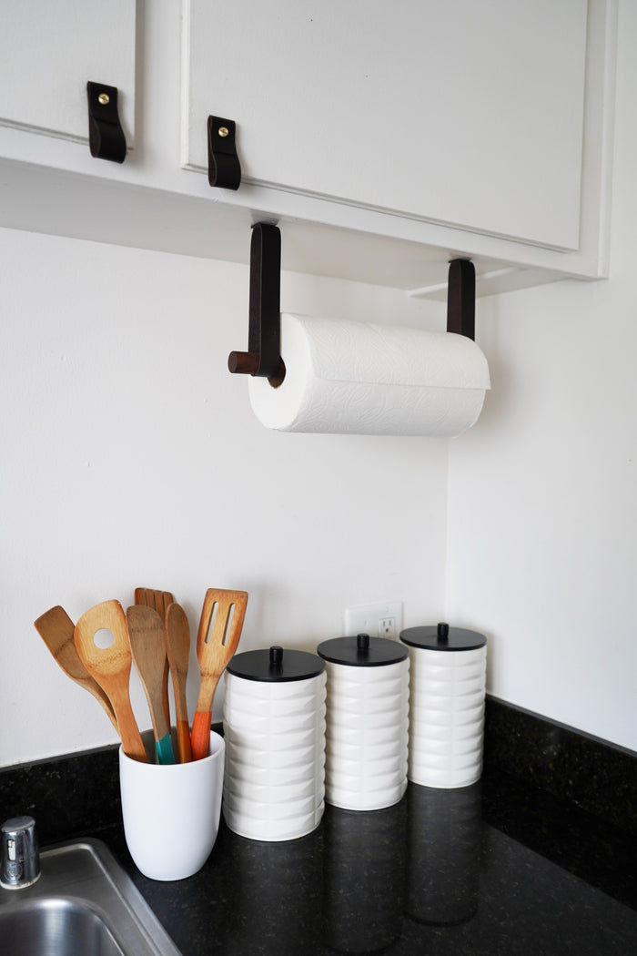 Wall-mounted paper towel holder installed under cabinet — leather strap and wood dowel kitchen organization system.