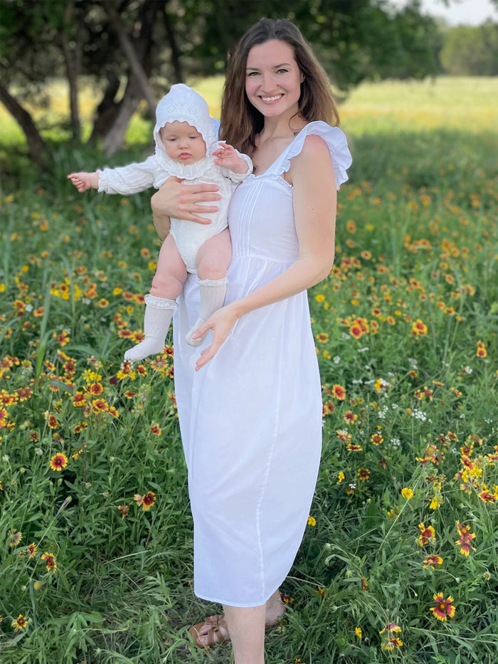 A woman standing in a field holding a baby wearing a white cotton nightgown with ruffle sleeves and decorative buttons.