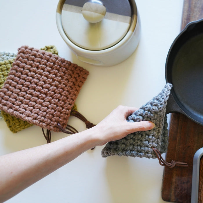 Set of woven cotton potholders with leather hanging loop displayed near stovetop — modern farmhouse kitchen accessory for oven use and heat protection.