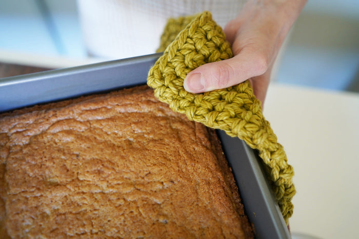 Olive green woven potholder placed under cast iron skillet — heat-resistant cotton hot pad with hanging loop for functional kitchen organization.