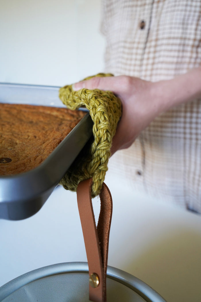 Keyaiira Woven cotton potholder with leather strap lifting hot baking dish — durable heat-resistant hot pad for oven-to-table serving.