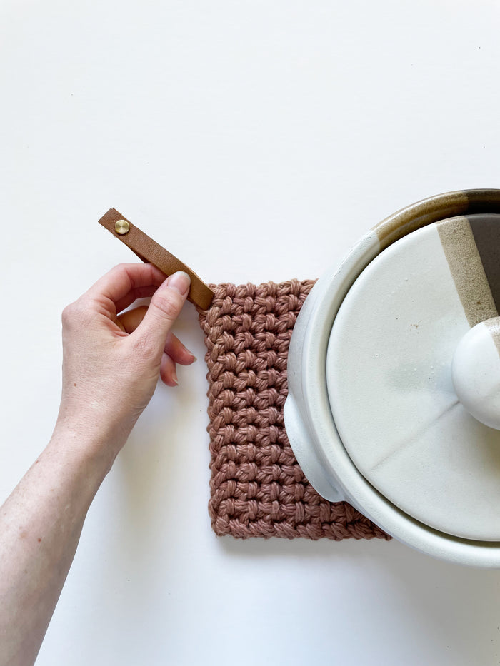 Blush-toned woven cotton potholder resting on pot lid with leather strap — modern handcrafted hot pad for stovetop and oven use.