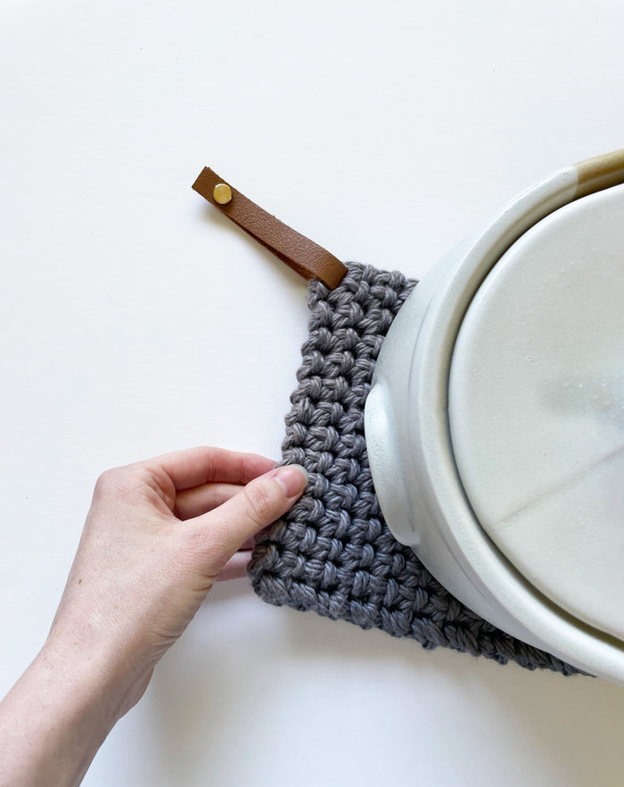 Neutral woven cotton potholder with leather strap hanger displayed under pot lid — textured heat-resistant kitchen accessory.