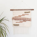 A hand woven wall hanging featuring light colors with mauve tones and rustic orange lines, displayed on a narrow slice of walnut wood.