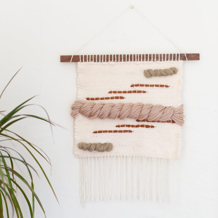 A hand woven wall hanging featuring light colors with mauve tones and rustic orange lines, displayed on a narrow slice of walnut wood.