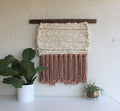Keyaiira handmade woven wall hanging with thick cream wool weaving and soft mauve fringe, mounted on a walnut wood dowel, showcasing modern textured fiber art styled with potted plants