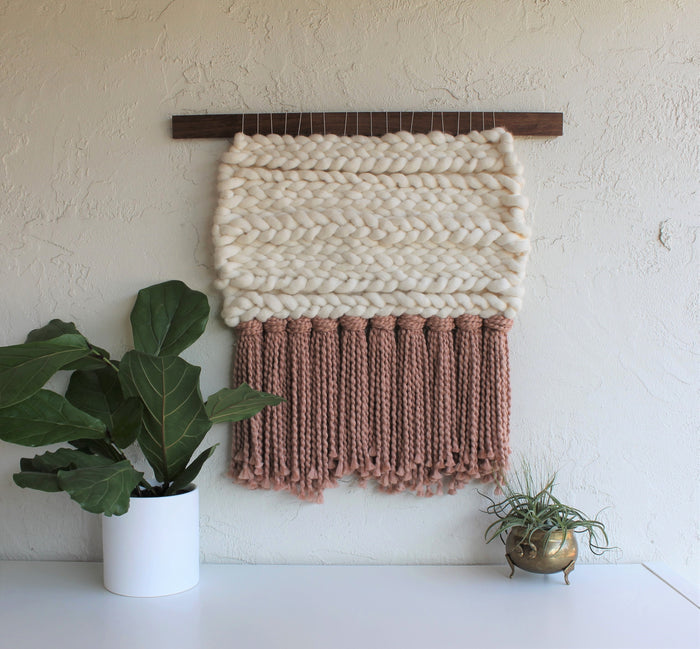 Keyaiira handmade woven wall hanging with thick cream wool weaving and soft mauve fringe, mounted on a walnut wood dowel, showcasing modern textured fiber art styled with potted plants