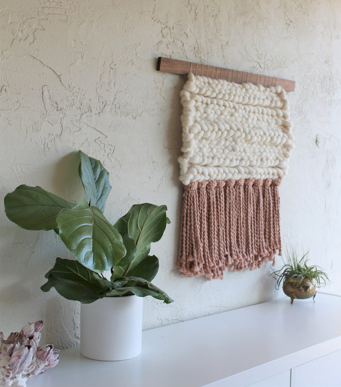 Keyaiira woven wall hanging styled on a white wall with greenery, showcasing thick braided cream wool and layered mauve fringe in modern minimalist decor