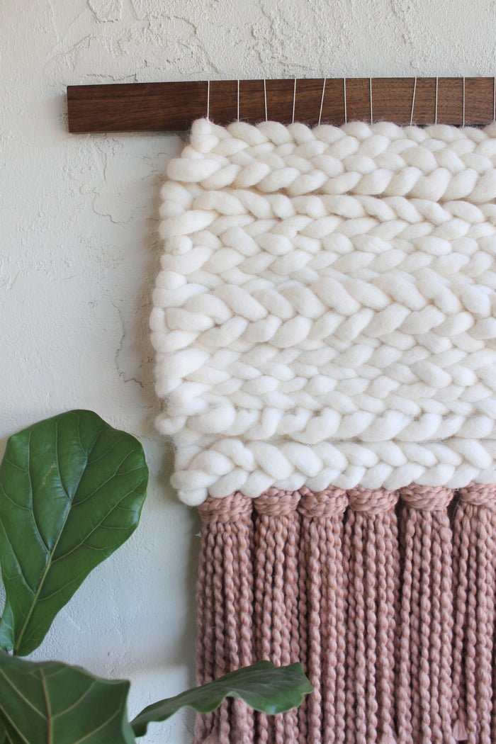 Close-up of Keyaiira fiber wall hanging showcasing chunky cream wool texture above soft mauve fringe, highlighting contrast and handcrafted weaving technique
