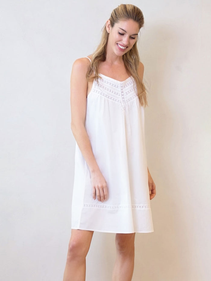 A woman standing in a white background wearing a white cotton nightgown with spaghetti straps and lace details.