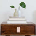 Keyaiira veg-tan wide leather pull mounted on a walnut nightstand drawer, shown with stacked books and ceramic vases, handcrafted modern furniture hardware for warm minimal interiors.