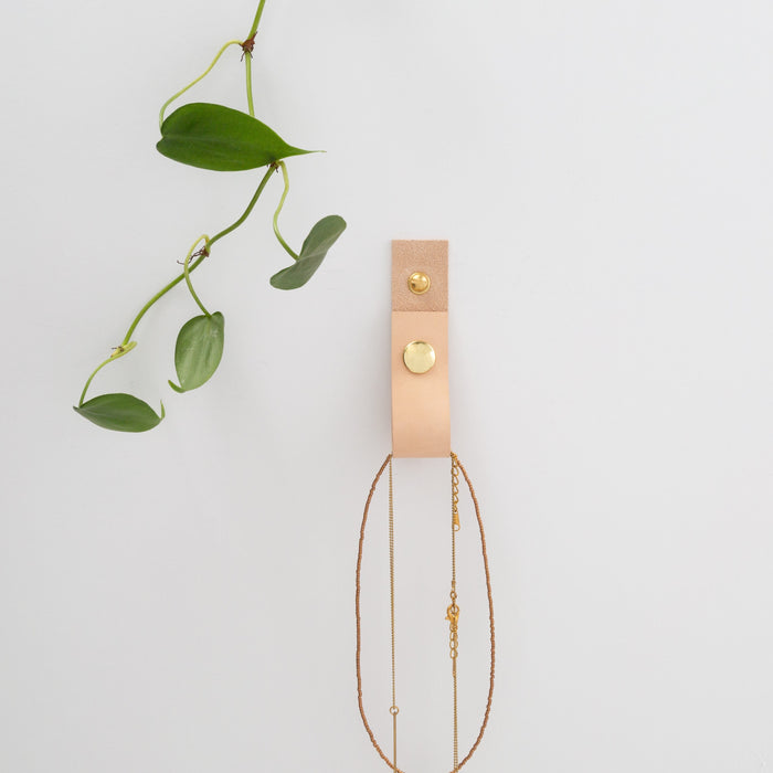Keyaiira natural vegetable-tanned leather wall strap with solid brass hardware mounted on white wall, styled as minimalist jewelry holder with delicate gold chains.