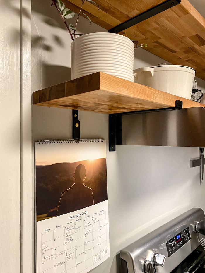 Keyaiira leather wall straps mounted beneath wood kitchen shelves holding hanging cookware — modern leather wall hooks with brass hardware for kitchen organization.