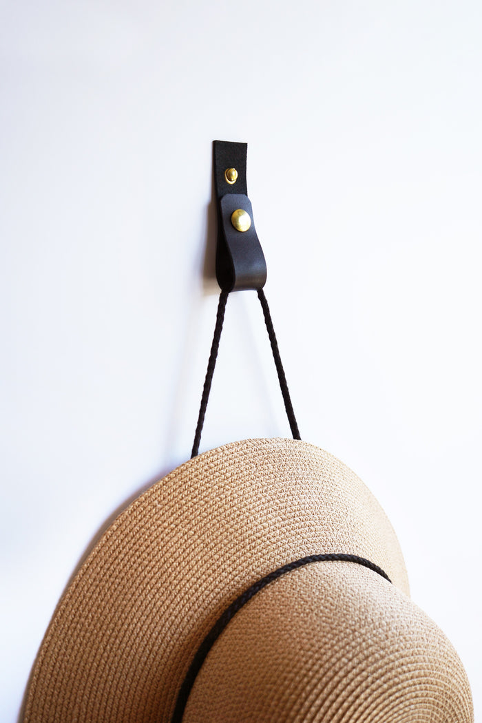 Keyaiira black leather wall strap with brass hardware holding woven hat — modern minimalist leather wall hook for entryway storage.