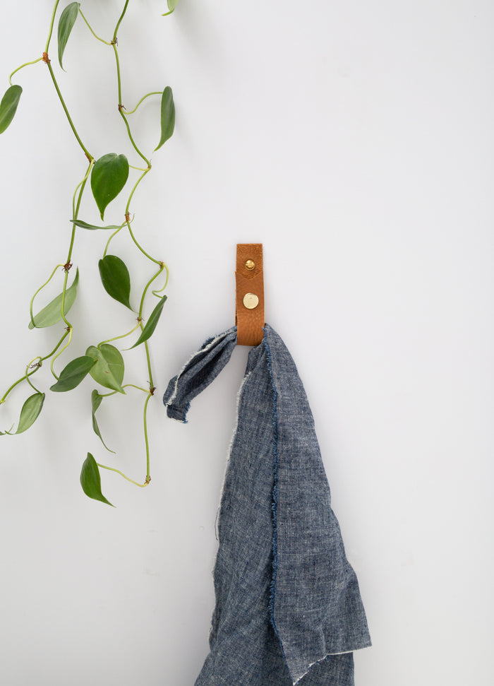 Keyaiira cognac leather wall strap mounted on white wall holding grey towel, styled with trailing greenery — handcrafted leather wall hook for bathroom or kitchen.