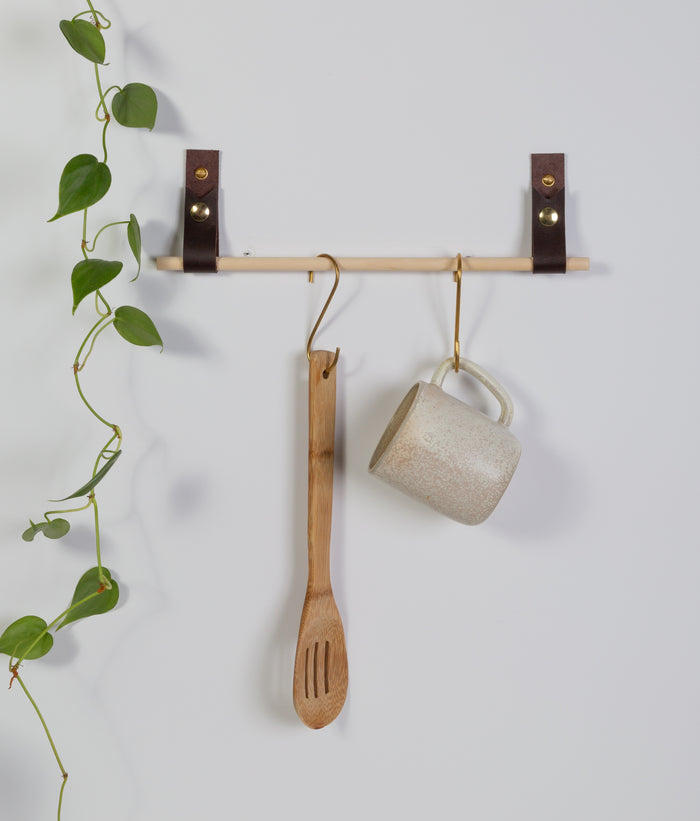 Two Keyaiira leather wall straps mounted with brass rod supporting hanging mug and wooden utensil — modern leather wall storage solution.