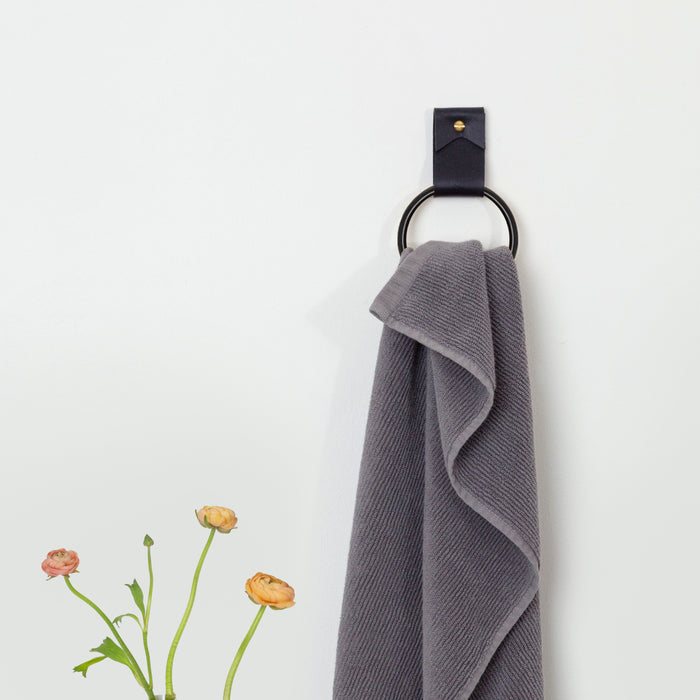 Small Wide Leather Wall Strap in black with brass hardware and ring minimalist handcrafted leather towel holder shown with gray towel. Modern functional wall hardware for bathroom or kitchen by Keyaiira.