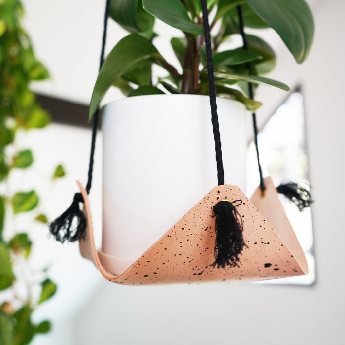 Keyaiira leather hanging planter with black cord and natural speckled leather cradle holding a white ceramic pot and trailing greenery. Handcrafted modern plant hanger for minimal interiors.
