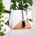 Keyaiira leather hanging planter with black cord and natural speckled leather cradle holding a white ceramic pot and trailing greenery. Handcrafted modern plant hanger for minimal interiors.