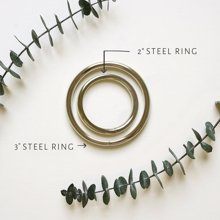 Two steel rings, one with a 2in diameter and a 3in diameter, displayed on a white background with green foliage in the background. The smaller ring is highlighted in an inset image showing a close-up of the surface and the text '3 STEEL RING' indicating the difference in size.