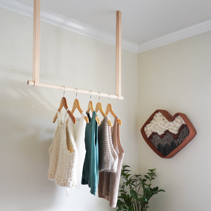 Minimal wooden hanging rail rack in a modern bedroom holding knit sweaters and tops  natural leather straps and birch wood dowel for functional, stylish clothing storage.