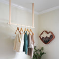 Minimal wooden hanging rail rack in a modern bedroom holding knit sweaters and tops  natural leather straps and birch wood dowel for functional, stylish clothing storage.