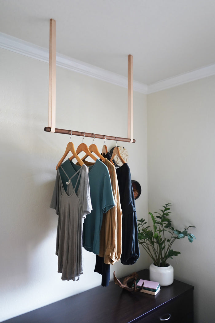 Ceiling-mounted wooden garment rack with vegetable tanned leather straps by Keyaiira, used for hanging clothes in a minimalist bedroom. Handcrafted modern home organization solution.