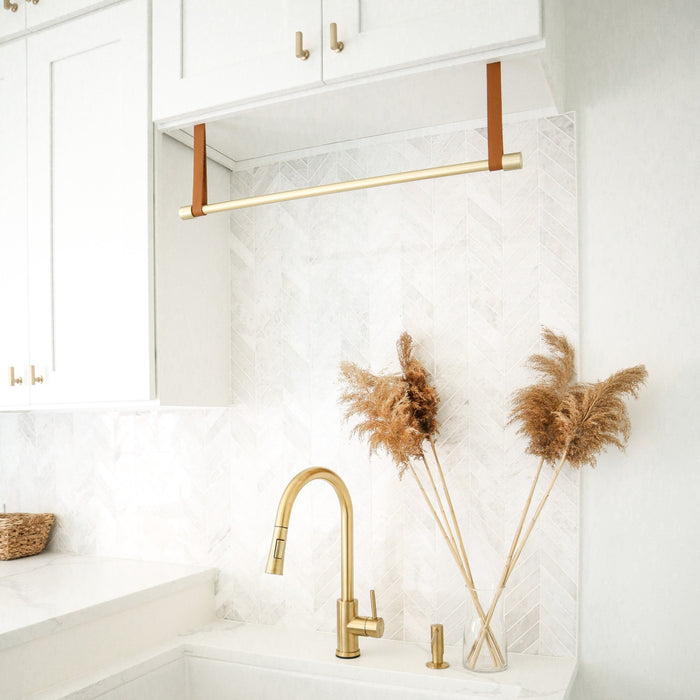 Keyaiira under-cabinet hanging rail with brass dowel and tan leather straps installed above a sink. Handcrafted modern home storage accent for kitchen or laundry decor.