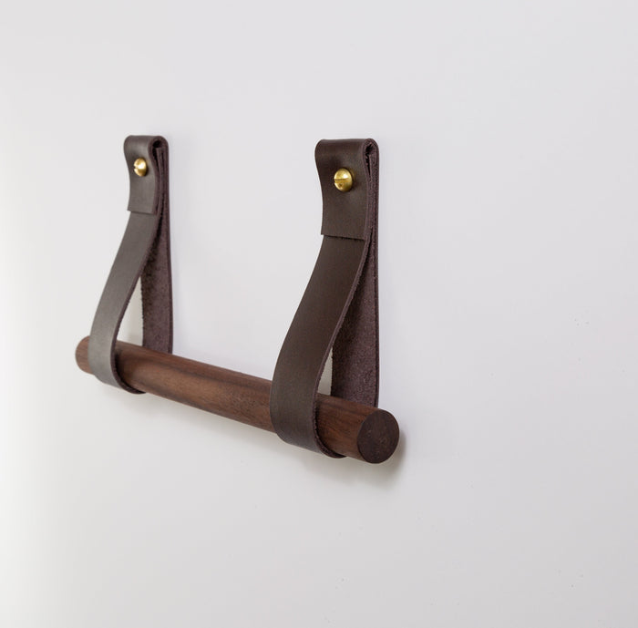 Mahogany brown leather toilet paper holder with walnut wood dowel and brass hardware mounted on a white wall.