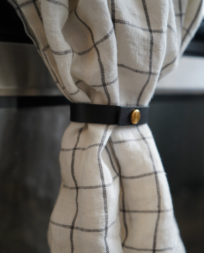 Keyaiira Towel Hugger leather loop in black shown holding a white check linen towel on a stainless steel appliance handle. Handcrafted vegetable-tanned leather with brass hardware for minimal functional kitchen decor.