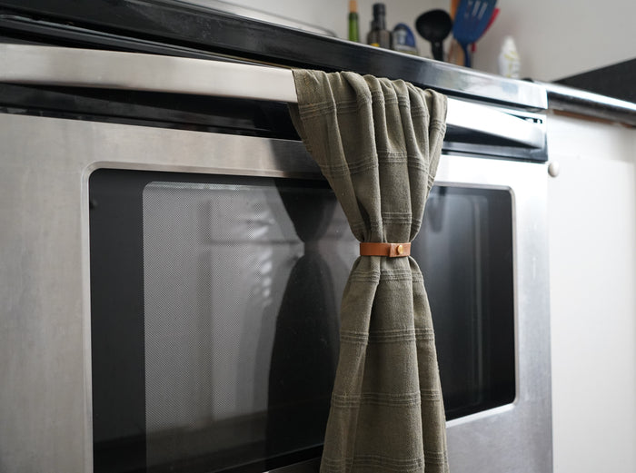 Keyaiira Nicotine colored leather Towel Hugger displayed in use holding a green waffle weave towel on a stainless steel oven. Sustainable handcrafted home accessory designed for functional, minimalist kitchen organization.