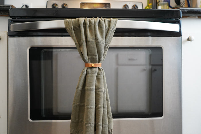 Keyaiira Nicotine leather Towel Hugger securing a green waffle-textured towel on a stainless steel oven handle. Minimal handcrafted kitchen decor designed for sustainable, functional everyday use.