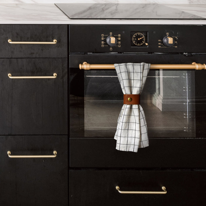 Keyaiira Wide Towel Hugger in warm cognac leather installed on brass oven handle, securing kitchen towel with solid brass hardware — modern farmhouse leather kitchen organizer.