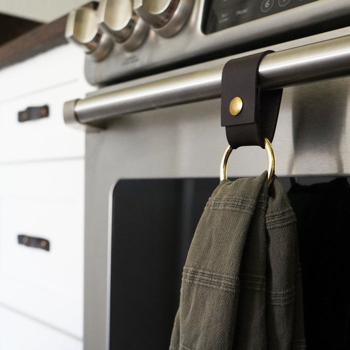 Keyaiira dark brown leather hanging strap installed on stainless steel oven handle with solid brass hardware ring holding kitchen towel — modern minimalist leather kitchen organizer.