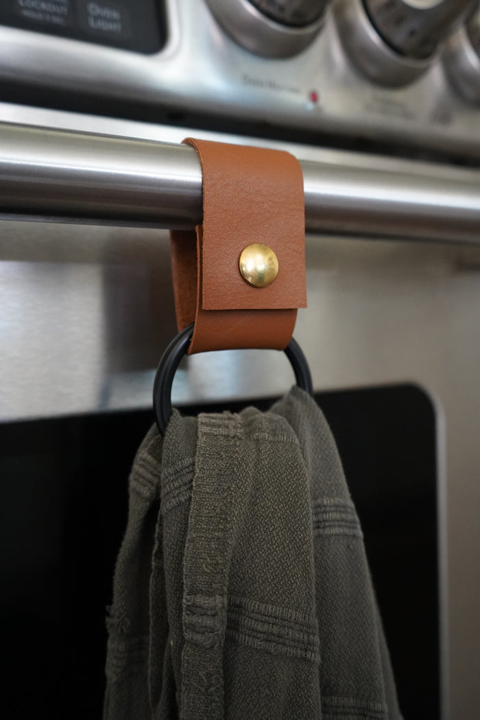 Keyaiira handcrafted leather towel ring with matte black ring mounted on stainless oven handle holding a green towel. Modern sustainable kitchen accessory made from byproduct leather.