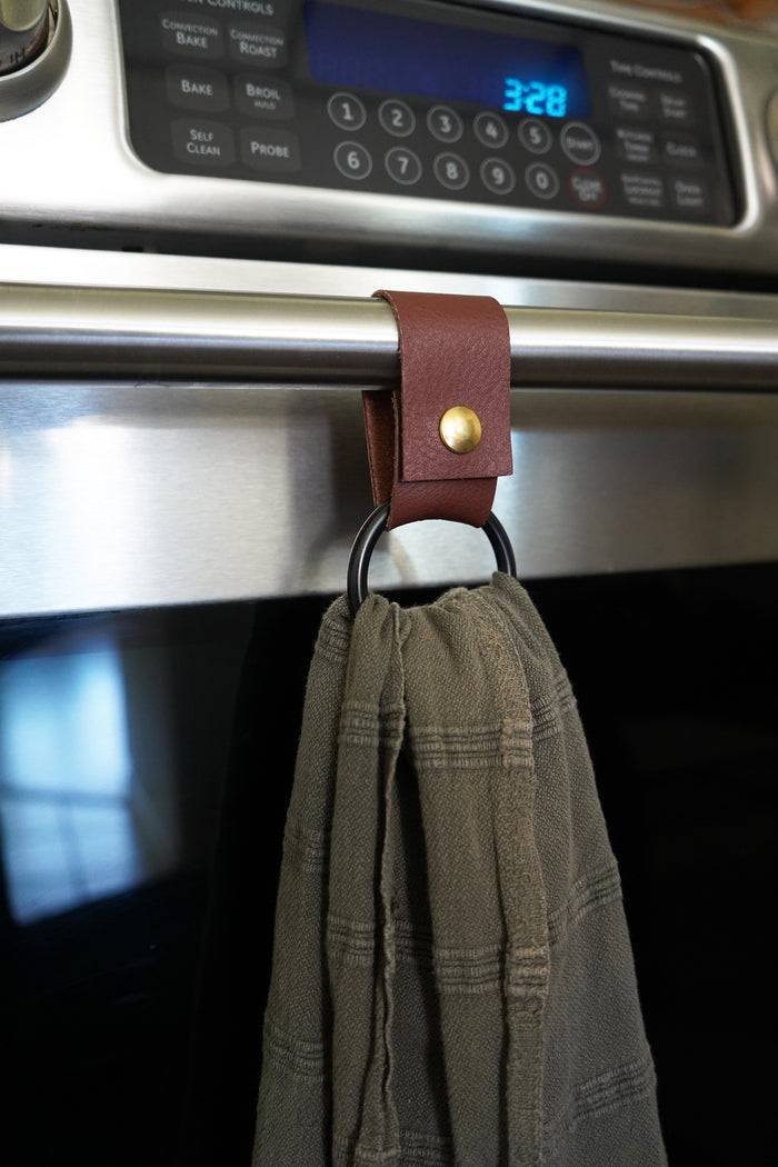 Keyaiira handcrafted towel ring in rich  leather with matte black ring  elegant, sustainable kitchen accessory shown on stainless oven handle.
