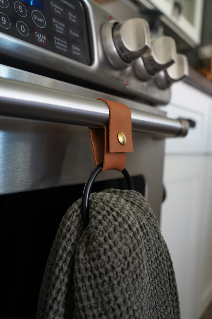 Keyaiira handcrafted leather towel ring in Nicotine with matte black ring shown hanging on a stainless steel oven handle sustainable leather kitchen accessory for modern functional decor.