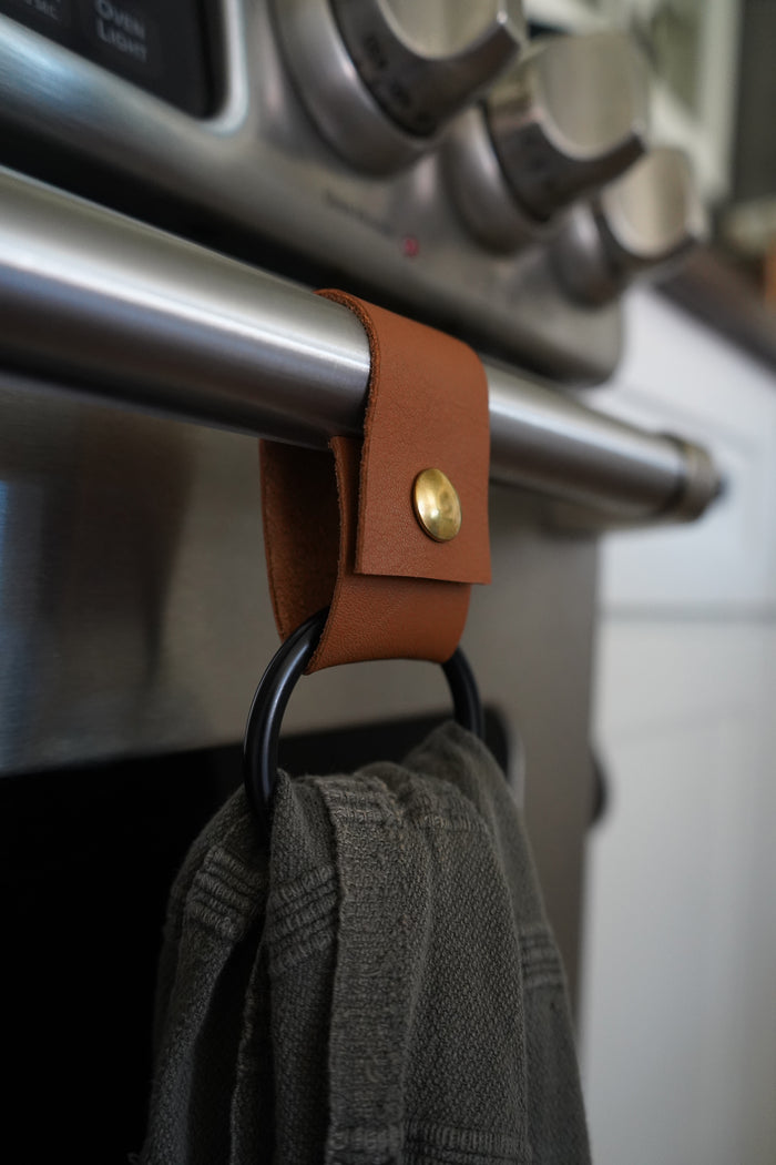 Keyaiira nicotine leather towel ring with matte black ring, Handcrafted sustainable leather kitchen accessory shown hanging on stainless steel oven handle.