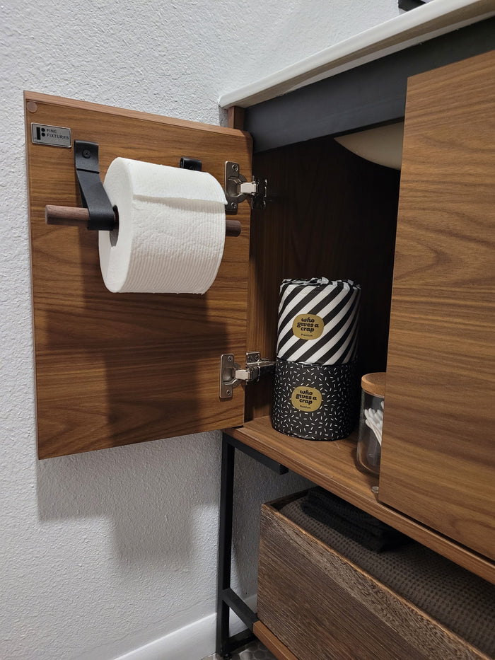 Keyaiira modern leather toilet paper holder in black leather shown on dark wood cabinetry. Handcrafted bathroom accessory combining warm natural materials and clean, functional design.