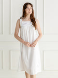 A woman wearing a white sleeveless nightgown with delicate embroidery on the chest, standing against a white paneled wall.